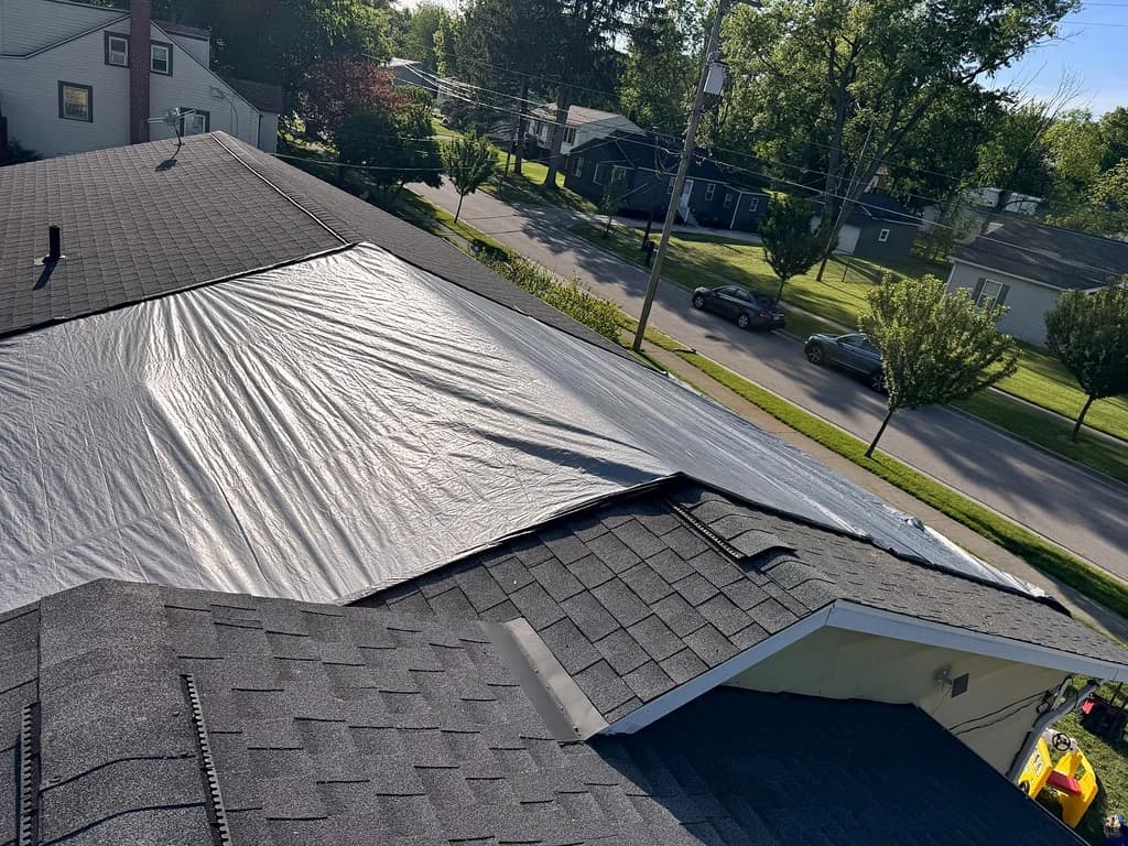 A tarp fitted over a damaged section of a shingle roof, with the street and neighboring homes below